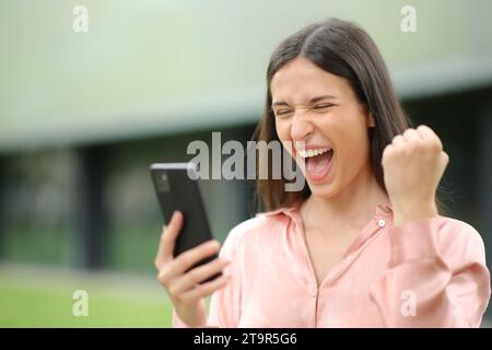 Donna elegante ed eccitata che festeggia il successo tenendo il telefono all'aperto Foto Stock