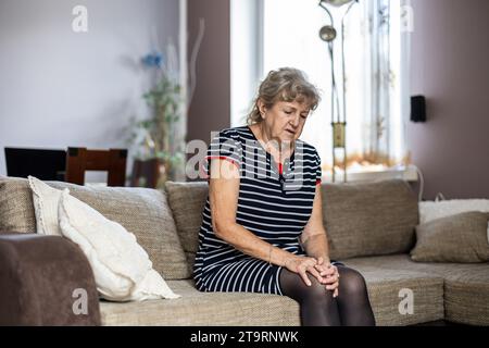 Donna anziana seduta sul divano a casa e affetta da dolore al ginocchio Foto Stock