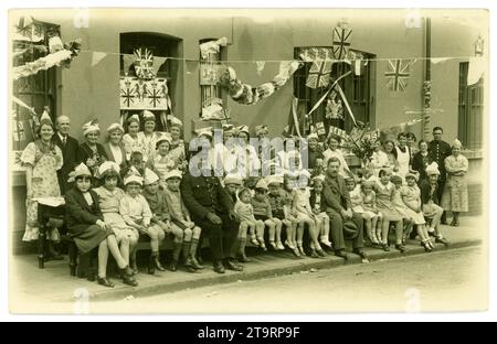 Cartolina originale degli anni '1930, affascinante immagine di bambini sorridenti che indossano cappelli da festa, che posano per una foto per strada con i genitori/adulti, prima di una festa del tè celebrativa, per celebrare l'incoronazione di George V1 (padre della regina Elisabetta), molte bandiere, bunting, banner, cappelli e personaggi da festa. Moda del tempo. Due poliziotti del Galles del Sud che si occupano per assicurarsi che non si eccitino tutti troppo. Eleanor Street, Tonypandy, Rhondda, South Wales, Regno Unito 1937. Foto Stock