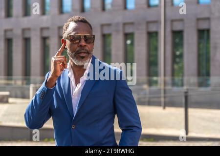 Un uomo d'affari africano maturo che indossa occhiali da sole e tuta che usa gli auricolari per parlare all'aperto Foto Stock