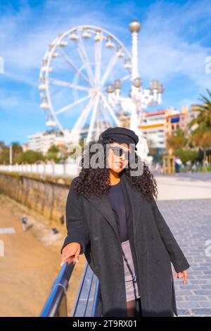 Ritratto di una donna latina alla moda in abiti invernali in piedi su una passeggiata accanto alla spiaggia in città Foto Stock