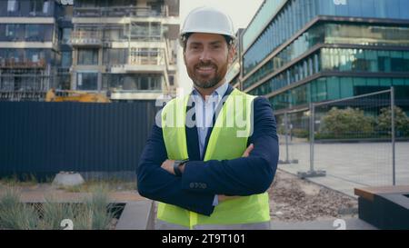 Sorridente uomo caucasico di mezza età lavoratore in hardhat felice sorriso ingegnere appaltatore in giubbotto da casco architetto ritrattista con le mani incrociate Foto Stock