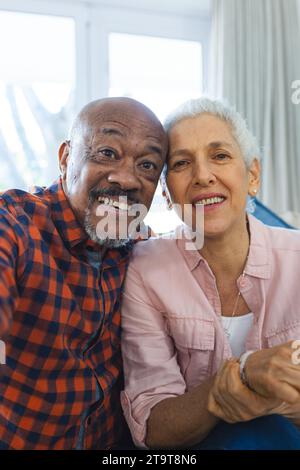 Coppia anziana felice e diversificata che effettua videochiamate nel soleggiato soggiorno. Stile di vita, pensionamento, stile di vita senior, unicità, comunicazione e domestico Foto Stock