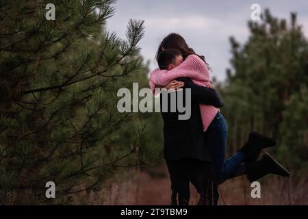 La coppia felice gira nella foresta di conifere lungo il sentiero. Il giovane abbraccia strettamente la donna dai capelli ricci con un maglione rosa. Fidanzamento invernale, matrimonio autunnale. Proposa Foto Stock