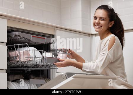 una donna attraente che scarica la tazza dalla lavastoviglie automatica aperta con utensili puliti all'interno in una moderna cucina bianca. Foto Stock