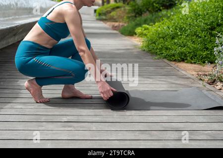 Primo piano di una donna che stende un tappetino per esercizi su una passerella in un parco al sole. Foto Stock