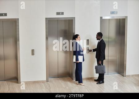 Veduta ad ampio angolo della donna d'affari caucasica con tazza e computer portatile che parla con l'allegro uomo d'affari afroamericano con telefono e valigetta vestiti in abiti in piedi vicino agli ascensori nella lobby, spazio fotocopie Foto Stock