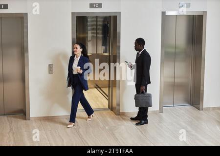 Ampia foto di un uomo d'affari afroamericano che tiene il telefono e la borsa che guarda la donna d'affari caucasica che esce dall'ascensore nell'edificio degli uffici Foto Stock