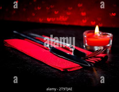 Tavolo di San Valentino con coltello, forchetta, candela a forma di cuore a fuoco rosso su sfondo bokeh con cuori rossi Foto Stock