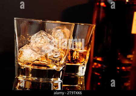 due bicchieri di whisky davanti alla bottiglia di whisky su un tavolo nero con riflessi e atmosfera calda Foto Stock