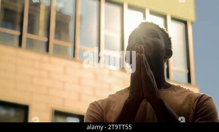 Concetto di religione uomo afro-americano che prega Dio per aiuto ragazzo di preghiera che implorano benedizione in città silhouette maschile al sole all'aperto alla luce del sole Foto Stock