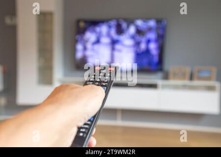 Primo piano con una mano maschile che tiene un controller TV nero con un flat tv sfocato sullo sfondo. Foto Stock