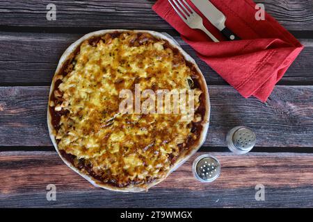 Pizza fresca fatta in casa servita su un tavolo di legno. Pizza di pollo con formaggio. Tovagliolo rosso, posate, coltello e forchetta, agitatori con sale e pepe. Vista dall'alto. Foto Stock