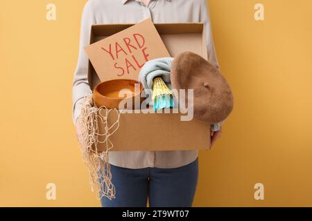 Donna che tiene una scatola di oggetti indesiderati e un cartello di cartone con testo IN VENDITA su sfondo giallo Foto Stock