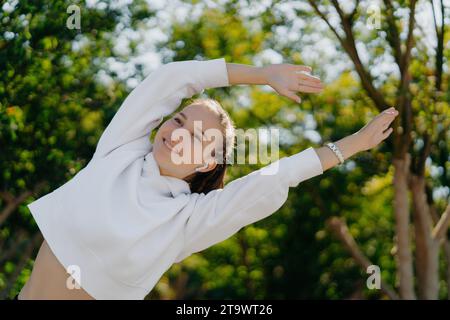 Giovane donna gioiosa che si allunga nel soleggiato parco, con gli auricolari accesi Foto Stock