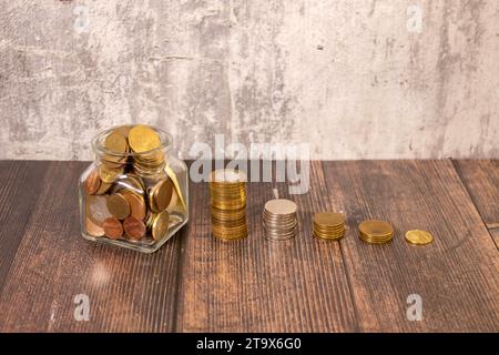 Monete in un vaso di vetro con il piccolo albero sulla parte superiore e la scala a monete sul tavolo. Concetto di risparmio economico e investimento finanziario in vaso di vetro. Scala a gettoni Foto Stock