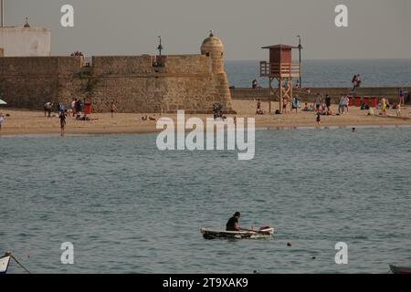 Un pescatore filtra la sua barca verso la spiaggia di Cadice con il Bastione di San Pedro dietro a Cadice, in Spagna. La grande spiaggia pubblica ospita pescatori, ristoranti e castelli. Foto Stock