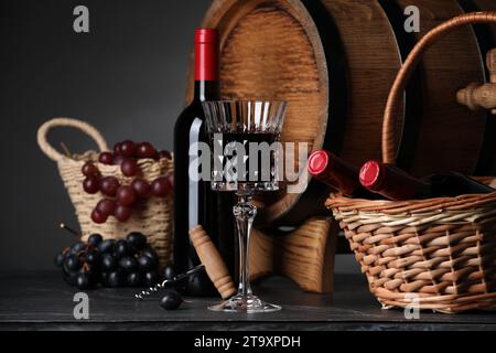 Bicchiere di vino rosso, bottiglie, cesto di vimini, uva e botte di legno su tavolo nero Foto Stock