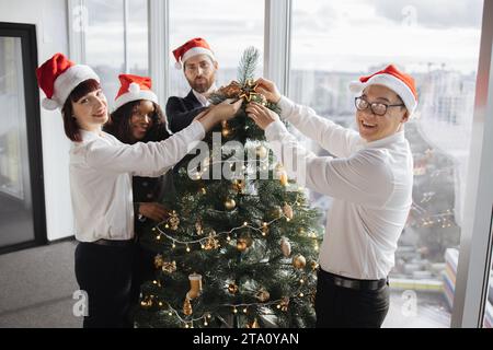 Team multietnico di persone che mettono l'arredamento dell'albero di natale in un ufficio festivo Foto Stock