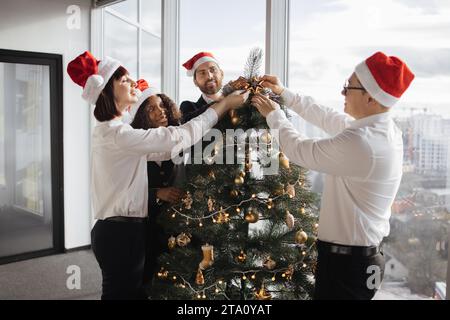 Team multietnico di persone che mettono l'arredamento dell'albero di natale in un ufficio festivo Foto Stock