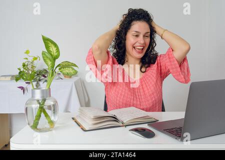 Giovane donna venezuelana latina con arricciacapelli e abiti casual rosa, è sorridente con le mani sulla testa seduta in ufficio a lavorare e a me Foto Stock