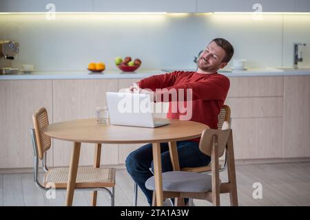 Uomo freelance positivo sorridente con gli occhi chiusi che si allunga seduto in cucina con un computer portatile a casa Foto Stock