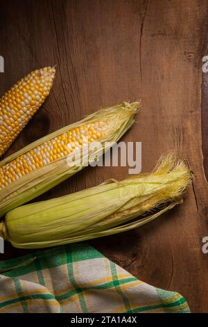 Vista ravvicinata di tre orecchie di mais dolce maturo con foglie verdi e asciugamano verde su fondo di legno vintage. Pannocchie con grani bianchi e gialli. Foto Stock