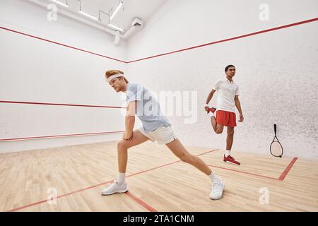 uomo sportivo a testa rossa con fascia che fa affondo vicino a un amico afro-americano all'interno del campo da squash Foto Stock
