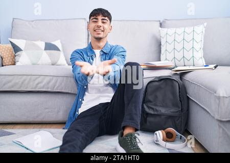 Giovane ispanico seduto sul pavimento a studiare per l'università sorridendo con le mani e i palmi insieme ricevendo o dando gesto. tenuta e protezione Foto Stock