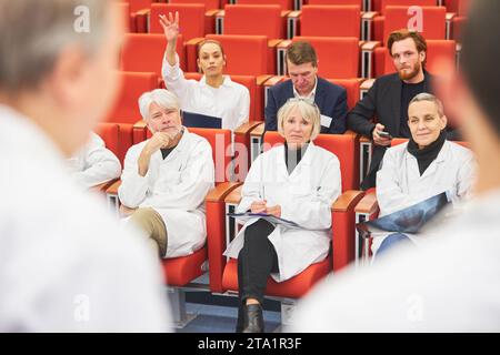 Medico femminile in udienza di conferenza medica facendo domanda all'oratore mentre seduto in sedia all'auditorium Foto Stock