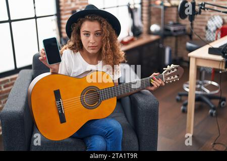 Giovane donna caucasica che suona la chitarra classica nello studio di musica, con smartphone rilassato con espressione seria sul viso. aspetto semplice e naturale Foto Stock