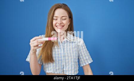 Giovane donna bionda sorridente sicura di tenere un test di gravidanza su sfondo blu isolato Foto Stock