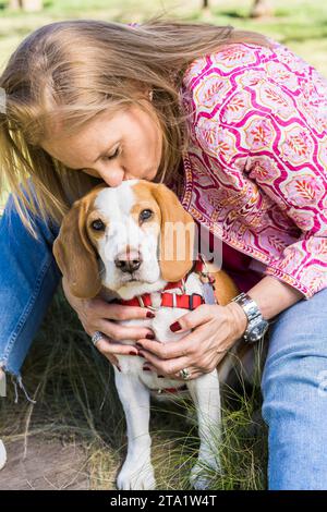 Bionda donna adulta che bacia il suo cane Beagle mentre guarda la macchina fotografica al parco Foto Stock