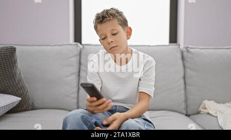 Adorabile ragazzino biondo che indossa un'espressione annoiata, che si affida comodamente sul divano a casa, sembra serio mentre si rilassa e guarda la tv in vita Foto Stock