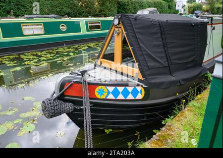 Corde nere legate alla prua di un narrowboat ormeggiato su un tranquillo ramo di canale tra le altre narrowboat e le erbacce galleggianti. Foto Stock