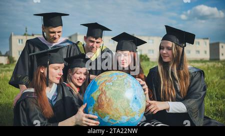 I laureati in abiti neri esaminano un globo geografico seduto sull'erba. Foto Stock