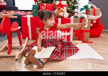 Adorabile ragazza cinese che punta di lato con un dito seduto sul pavimento vicino all'albero di natale a casa Foto Stock