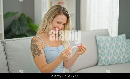 Giovane donna bionda sorridente sicura di tenere il test di gravidanza a casa Foto Stock