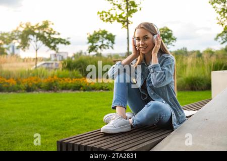 Una donna bionda siede su una panchina con le gambe incrociate, godendosi il tramonto nel parco attraverso le cuffie Foto Stock
