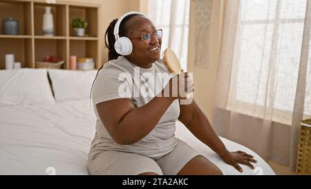 Donna afroamericana sicura di sé che si gode la mattina, si rilassa sul letto cantando una canzone usando una spazzola come microfono, ascoltando la sua voce attraverso la testa Foto Stock