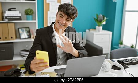 Scopri il nostro giovane lavoratore ispanico, adolescente, che si mette a dura prova con la sua videochiamata con lo smartphone mentre è in ufficio. il nostro pro dietro la sc Foto Stock