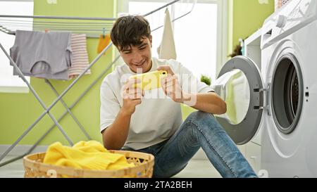 Bellissimo giovane adolescente ispanico che gioca con gioia a un videogioco mentre attende pazientemente il ciclo di lavaggio sui suoi vestiti nella vivace lavanderia di casa Foto Stock