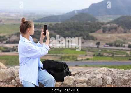 Vista posteriore di una giovane donna viaggiatrice con zaino si rilassa sulla collina, scatta foto delle montagne e del paesaggio della valle con lo smartphone. Zaino in spalla, AC Foto Stock