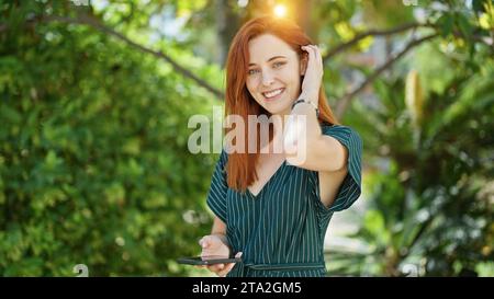 Giovane donna rossa che usa lo smartphone per pettinare i capelli al parco Foto Stock