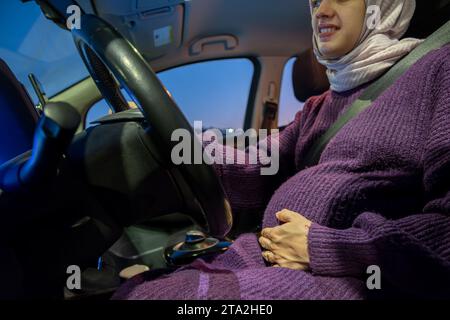 Donna musulmana araba incinta che guida un'auto di notte indossando abiti invernali, hijab e cintura di sicurezza, guidando in modo sicuro e sicuro Foto Stock