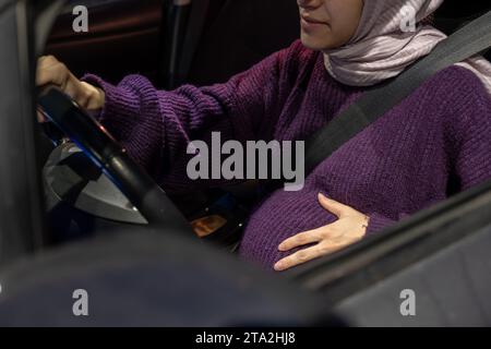 Donna musulmana araba incinta che guida un'auto di notte indossando abiti invernali, hijab e cintura di sicurezza, guidando in modo sicuro e sicuro Foto Stock