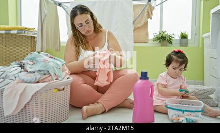 Madre e figlia condividono un momento serio mentre tengono insieme vestiti e un cestino di vimini nell'accogliente lavanderia della famiglia, un angolo della loro vigilia Foto Stock