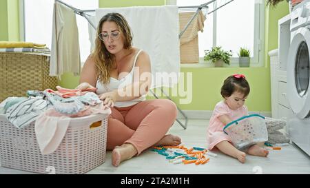 Madre e figlia condividono un momento serio mentre tengono insieme vestiti e un cestino di vimini nell'accogliente lavanderia della famiglia, un angolo della loro vigilia Foto Stock