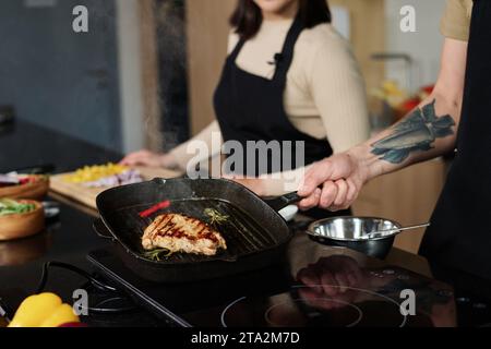 Primo piano di un uomo irriconoscibile che tiene una padella con un pezzo di carne preparato che lo mostra alla fotocamera con una donna sconosciuta in piedi sullo sfondo, concentrati sulla carne Foto Stock