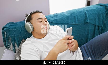 Bellissimo giovane cinese si è rilassato a casa, impegnato a guardare un video sul suo smartphone mentre si siede sul suo comodo divano, totalmente immerso nel Foto Stock